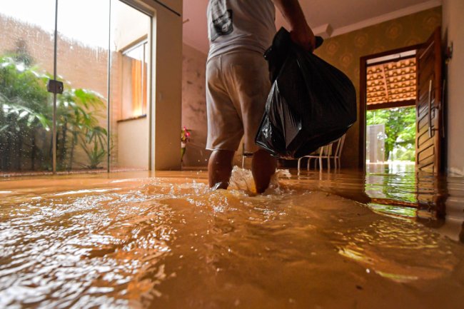 Chuvas na Bahia e em Minas Gerais: saiba como ajudar as vítimas Foto de um senhor, de costas, carregando um saco de lixo. Ele está na sala de casa e a água da enchente chega até os tornozelos