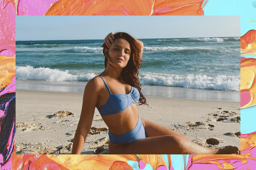 Larissa Manoela sentada em areia da praia com biquíni azul. O fundo da montagem tem tintas laranja, azul e rosa.