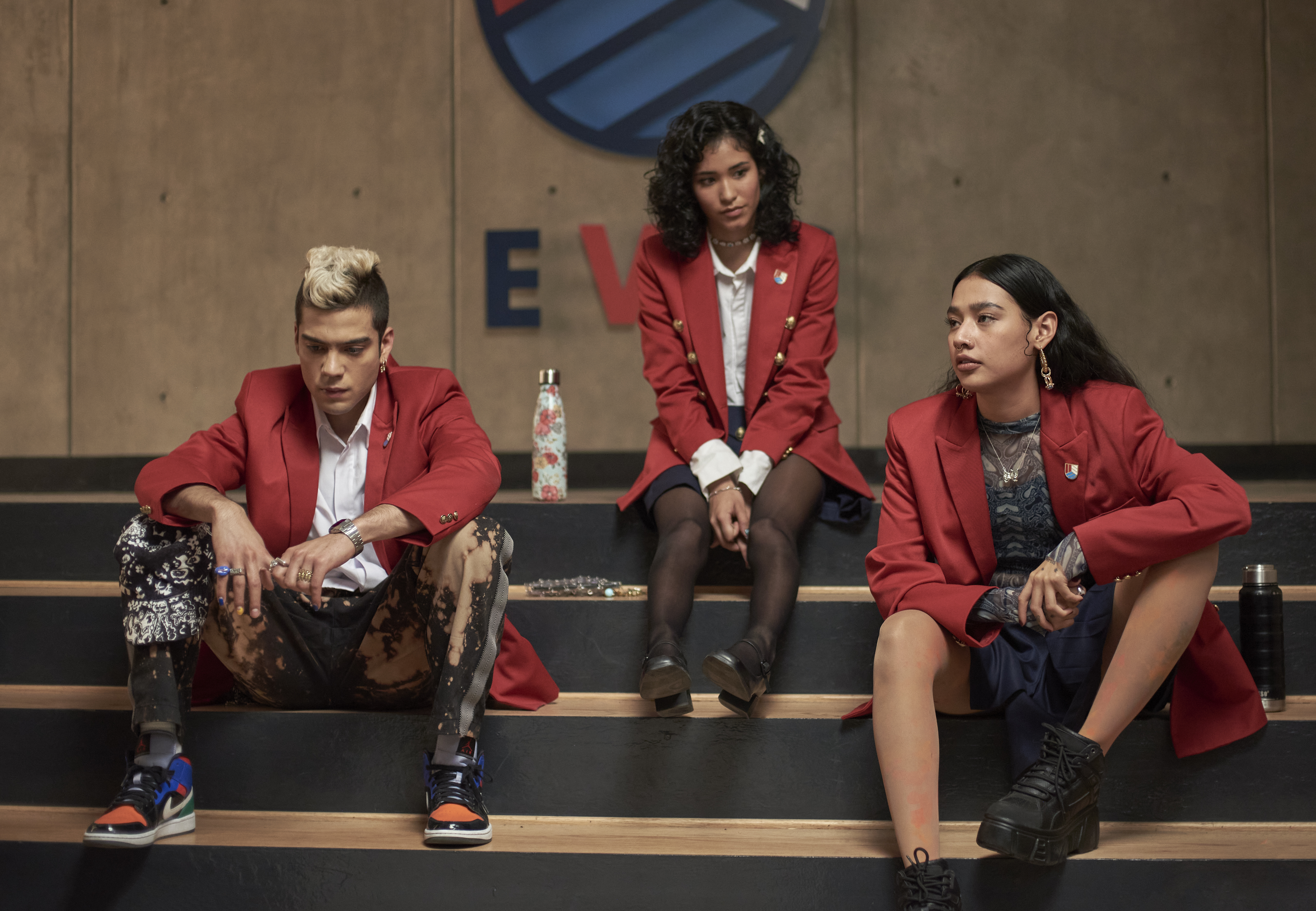 Os personagens Dixon, MJ e Andi, da série Rebelde, da Netflix, sentados em escada da escola