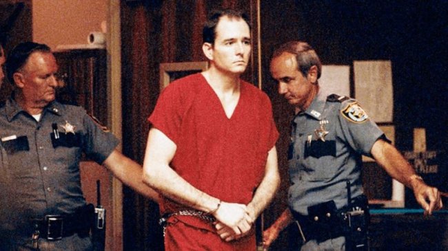 Foto do serial Killer Danny Rolling. Ele está algemado, com uma roupa vermelha e cercado por policias no tribunal