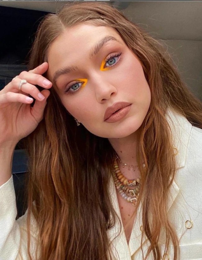 Foto da Gigi Hadid. Ela usa uma camisa branca, mix de colares, cabelo solto e maquiagem com batom nude e sombra laranja neon no canto interno dos olhos. Ela está com a mão direita na cabeça, olha para a câmera e não sorri.