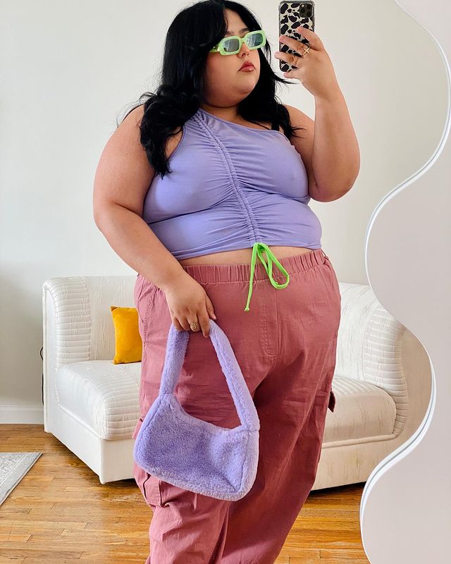 Selfie no espelho de uma mulher. Ela usa um top cropped lilás com detalhe franzido, calça rosa, bolsa baguete de pelúcia lilás e óculos de sol. Ela olha para o celular e não sorri.