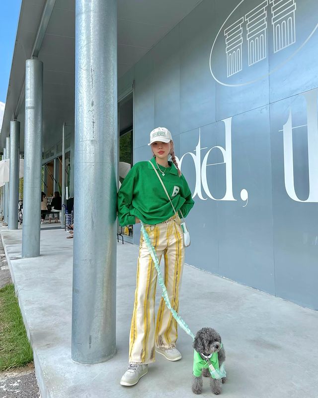Foto de uma mulher em uma calçada. Ela usa um boné branco, bolsa branca, moletom Verde Bottega, calça amarela e tênis branco. Ela segura a coleira de um cachorro com a mão direita, olha para a câmera e sorri levemente.