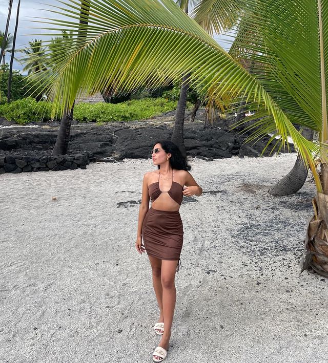 look-conjunto-saia-cropped Foto de uma mulher na praia. Ela usa um top cropped marrom de amarração, saia marrom com detalhe franzido nas laterais, sandália branca e óculos de sol. Ela olha para o lado e não sorri.