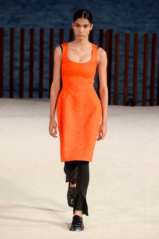 look-vestido-e-calca Foto de uma mulher na passarela. Ela usa um vestido laranja, calça preta e mocassim preto.