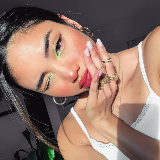 Selfie de uma mulher. Ela usa uma blusa de alcinha branca, cabelo preso, brinco de argola e maquiagem simples com detalhe neon no canto interno dos olhos.