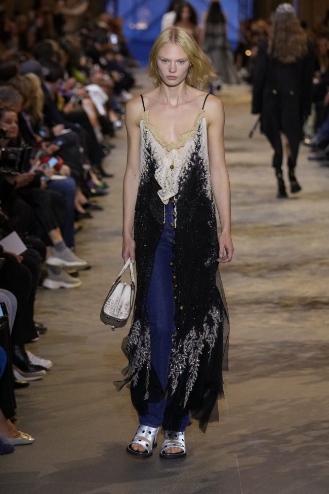 passerela-vestido-com-calca Foto de uma mulher desfilando em uma passarela. Ela usa um vestido de alcinhas longo azul com uma calça jeans com bolsa e sandália metalizada.
