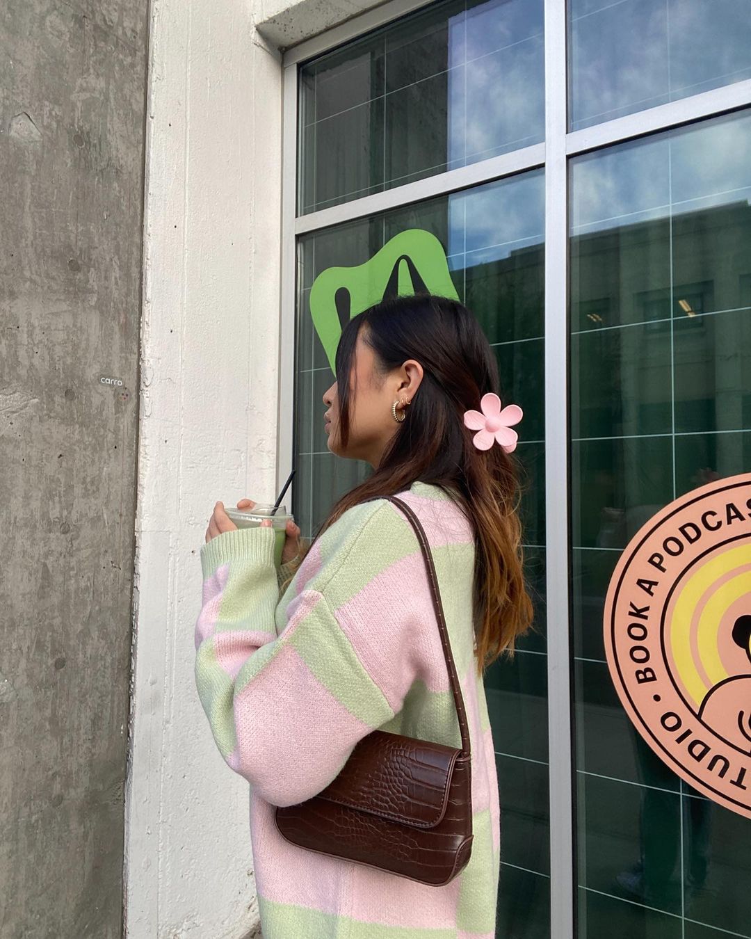 Garota de lado usando cardigan rosa e verde e bolsa marrom pendurada no ombro. Seu cabelo está preso com uma piranha em formato de flor rosa.