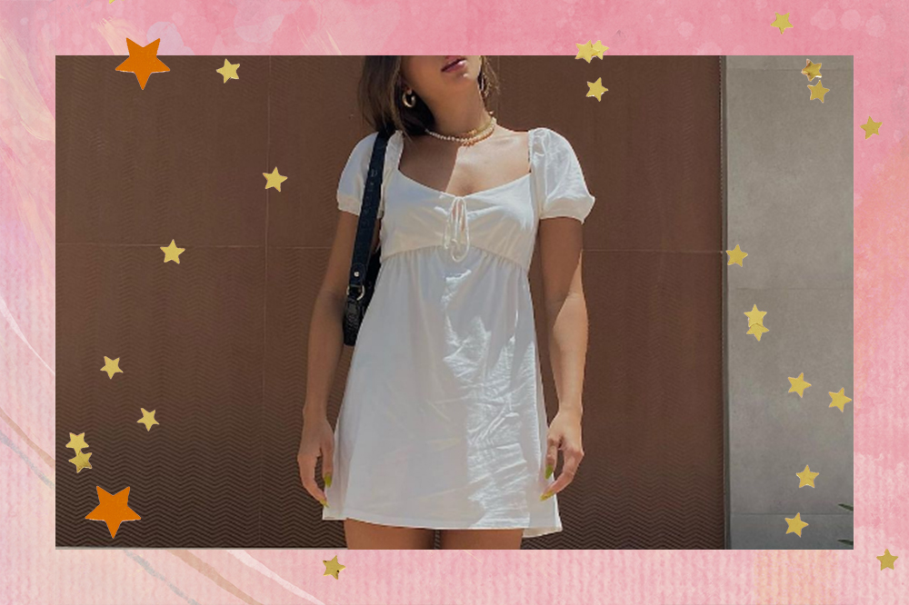 Garota usando vestido branco com mangas bufantes e uma bolsa preta pendurada no ombro. A montagem possui fundo rosa com estrelinhas douradas e laranjas