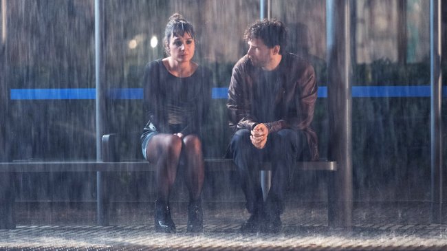 Foto de um casal sentado em um ponto de ônibus. Está chovendo muito, eles estão molhados e tristes um com o outro