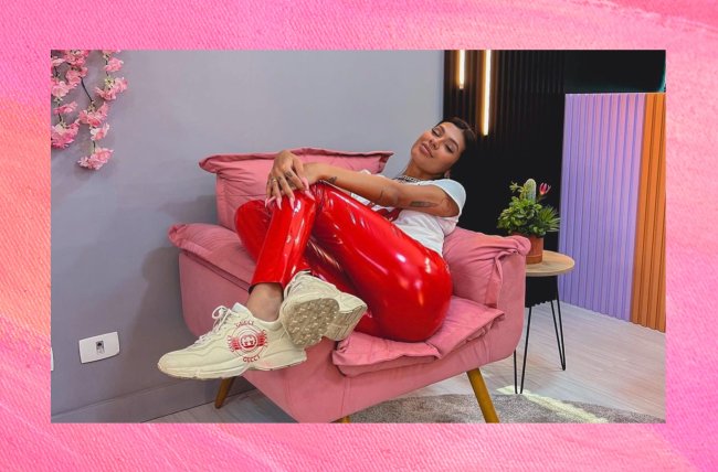 Pocah tem cabelos preos e médios, está vestida com uma calça vermelha, camiseta branca e está sentada em uma poltrona rosa. A sala tem paredes cinzas. A foto está centralizada e uma montagem com fundo rosa.