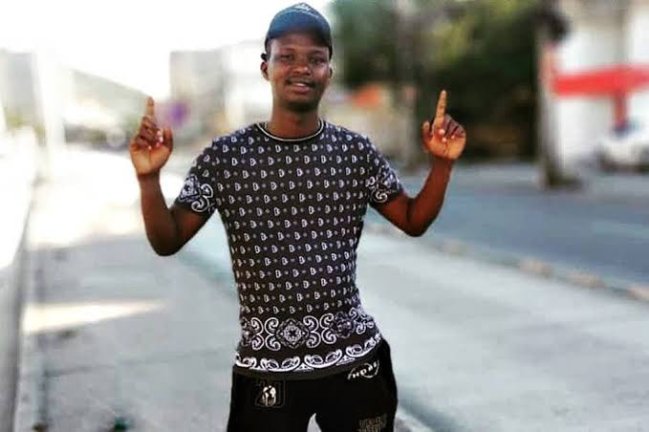Foto do Moïse mugenyi kabagambe, na rua. Ele está com uma calça preta, uma camiseta preta com estampas brancas. Está de pé com os braços levantados. Ele é um jovem negro.