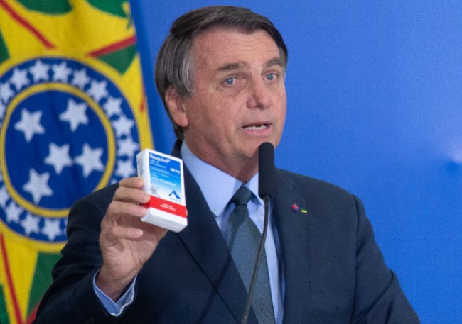 Presidente Bolsonaro em um fundo azul, com a bandeira nacional ao fundo. Ele está de terno, sem máscara, falando ao microfone e segurando uma caixa de remédio.