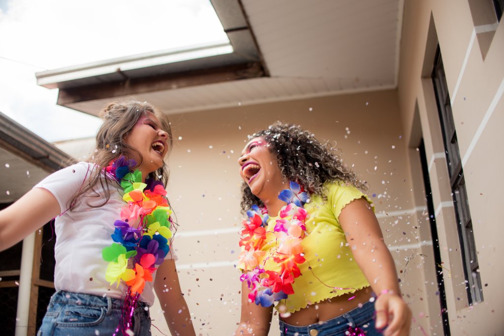 Duas meninas rindo muito com colares de Carnaval coloridos e em meio a confetes e purpurinas