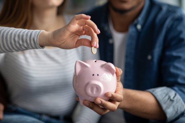 Foto de duas pessoas adultas segurando um cofrinho rosa de porco enquanto uma adolescente deposita uma moeda dentro dele. Não é possível ver os rostos.