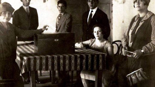 Foto em preto e branco de Celina Guimarães se alistando para ter o direito de votar. Ela está sentada a uma mesa, escrevendo em um pedaço de papel