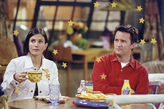 Fotos da Monica e do Chandler, de Friends, tomando café em uma mesa com comidinhas. Ela veste uma jaqueta branca e ele uma blusa vermelha.