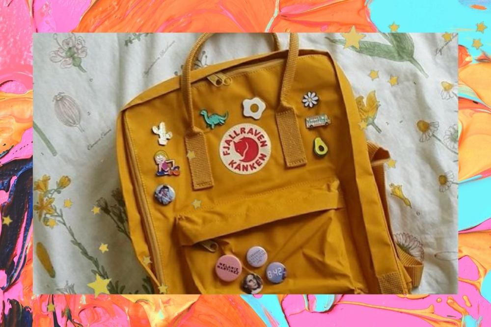 Montagem com o fundo colorido e detalhe de estrelas nas bordas com a foto de uma mochila amarela decorada com bottons em cima de uma cama.