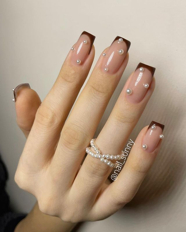 Foto de uma mão com as unhas decoradas com francesinha marrom e aplicação de pérolas.