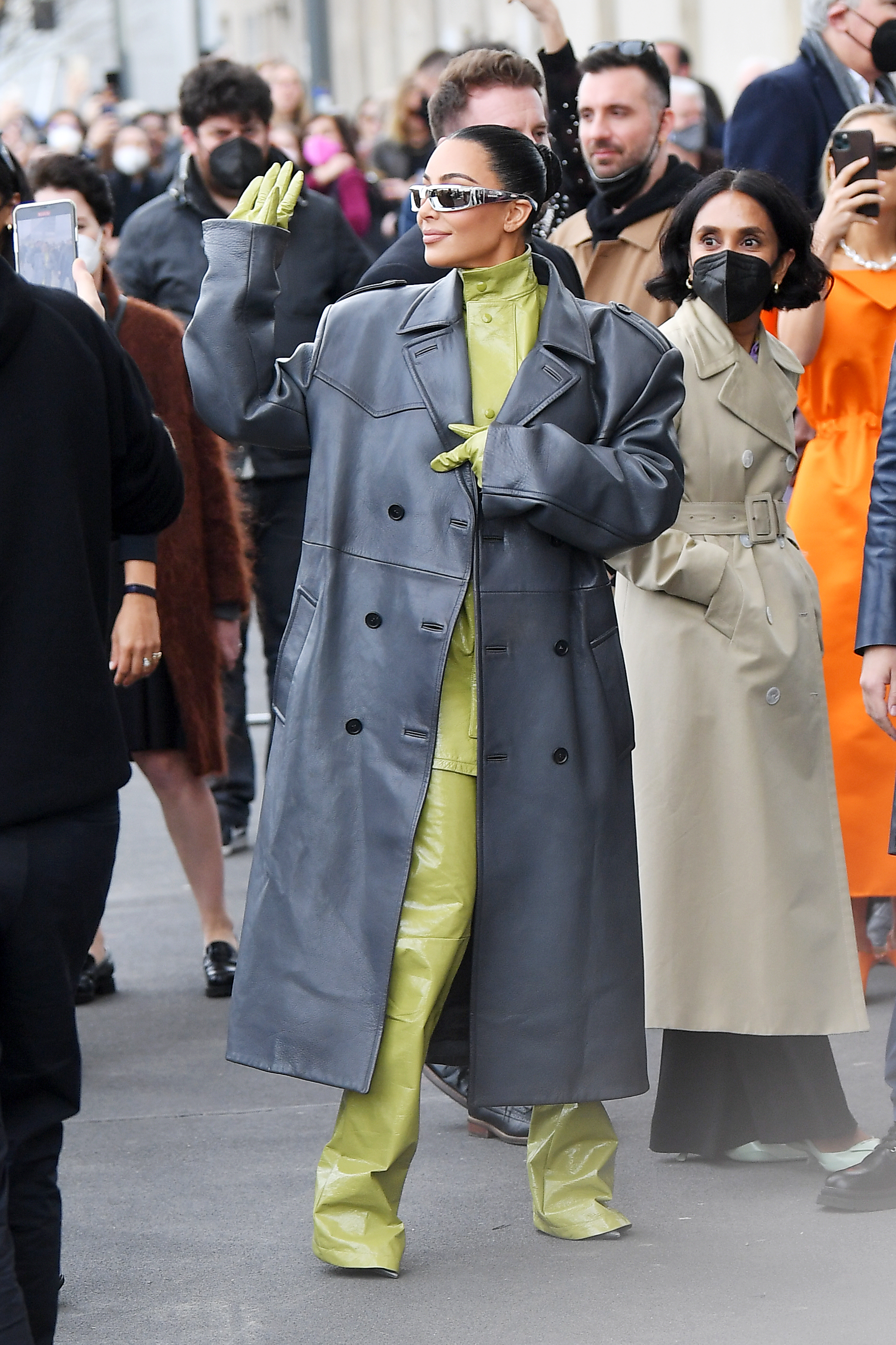Kim Kardashian na semana de moda de Milão usando macacão verde com sobretudo cinza e óculos escuros. Ela olha para o lado e levanta a mão para cumprimentar fãs