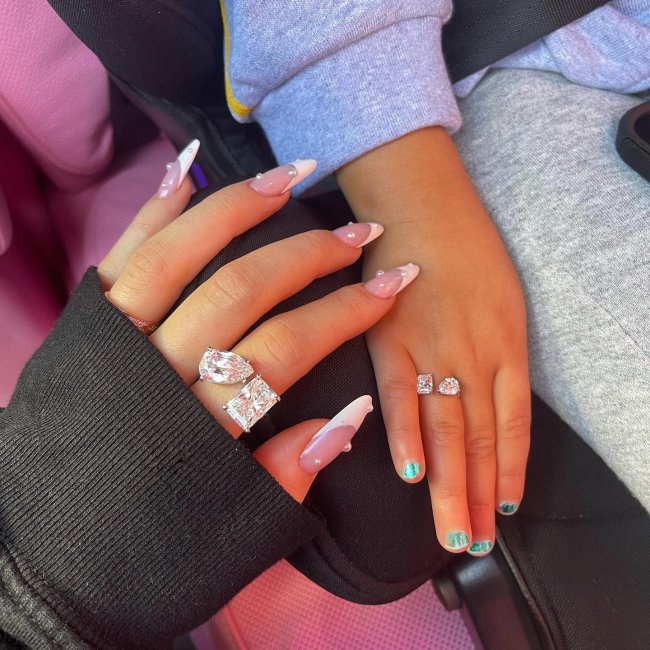 Foto das mãos da Kylie Jenner e sua filha Stormi. A Kylie está com as unhas longas com uma decoração de francesinha branca e pérolas e ambas usam anéis combinando.