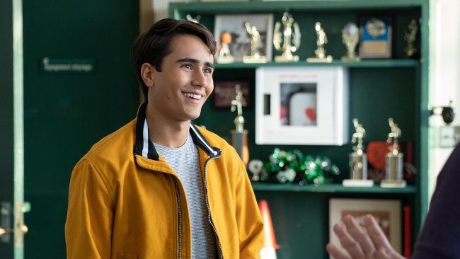 Cena de Love, Victor em que o personagem principal está sorrindo e usando um casaco amarelo com blusa branca; ao fundo é possível ver um móvel grande e verde com trófeus dourados