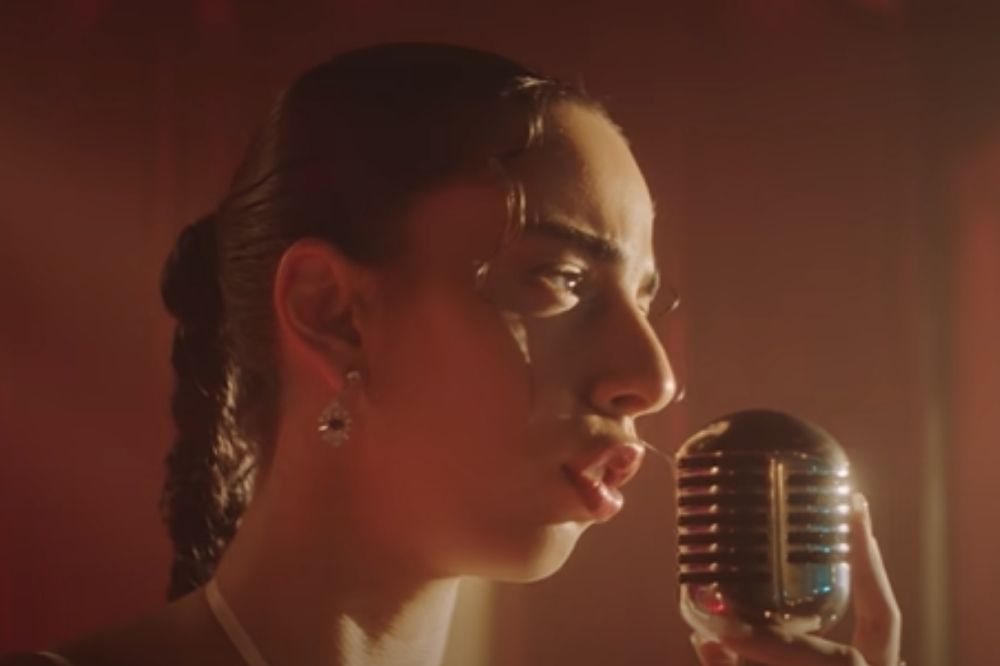 Marina Sena no clipe de Por Supuesto; ela está de perfil com o cabelo presto com uma trança e canta em um microfone retrô/vintage