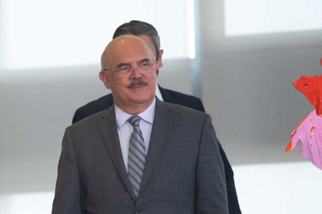 Foto de Milton Ribeiro, ministro da Educação, andando num grande corredor branco. Ele é um homem branco, de cerca de 60 anos, usa óculos, tem bigode, é careca e veste um terno cinza