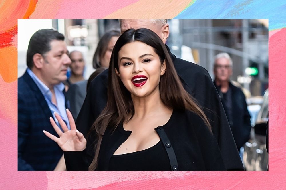 Selena com roupa preta e batom vermelha andando na rua e acenando.