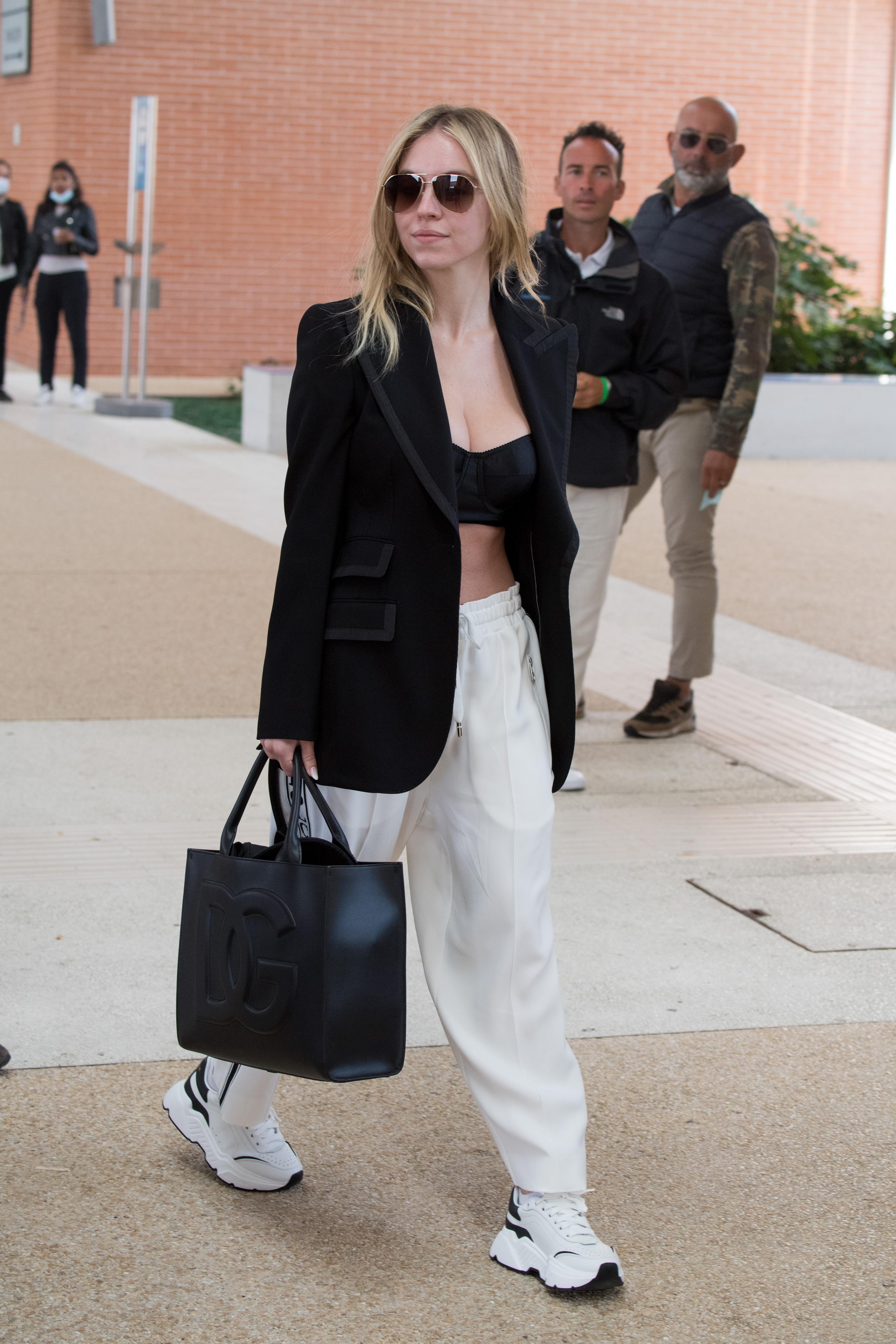 Sydney Sweeney andando na rua e usando look com blazer e top cropped pretos, calça e tênis brancos e segurando uma maxibolsa preta, além de estar usando óculos de sol