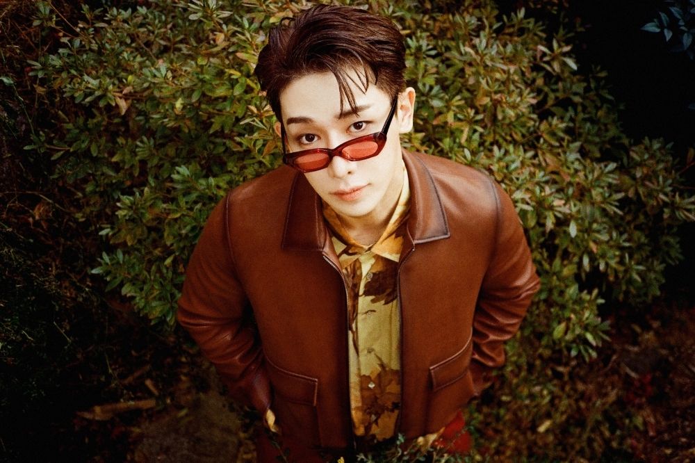 Wonho com jaqueta marrom e óculos de lente colorida em um jardim.