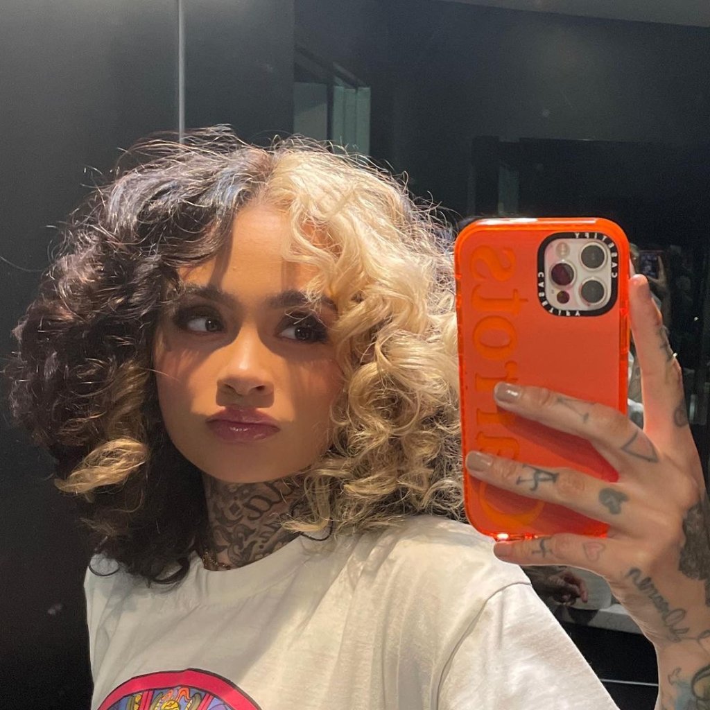 Selfie no espelho da cantora Kehlani; ela está com expressão neutra posando para foto enquanto segura o celular com capinha laranja e olha para a tela; ela usa uma camiseta branca e o cabelo está divido ao meio em duas cores, sendo metade preto e metade loiro
