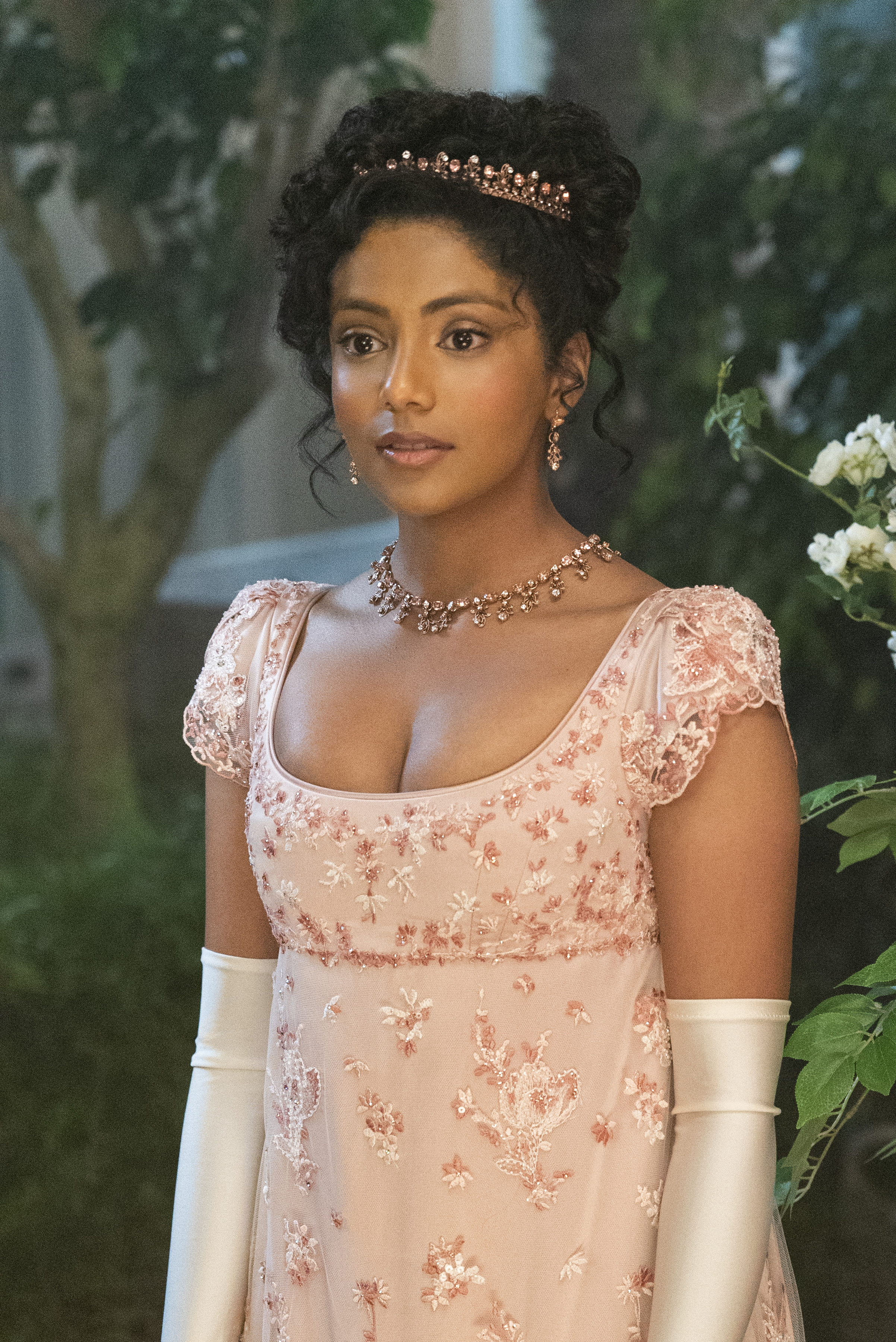 Imagem mostra Charithra Chandran caracterizada como Edwina Sharma usando joias de ouro rosa e vestido bordado com os mesmo elementos e luvas brancas em cena do 1º episódio da segunda temporada de Bridgerton.