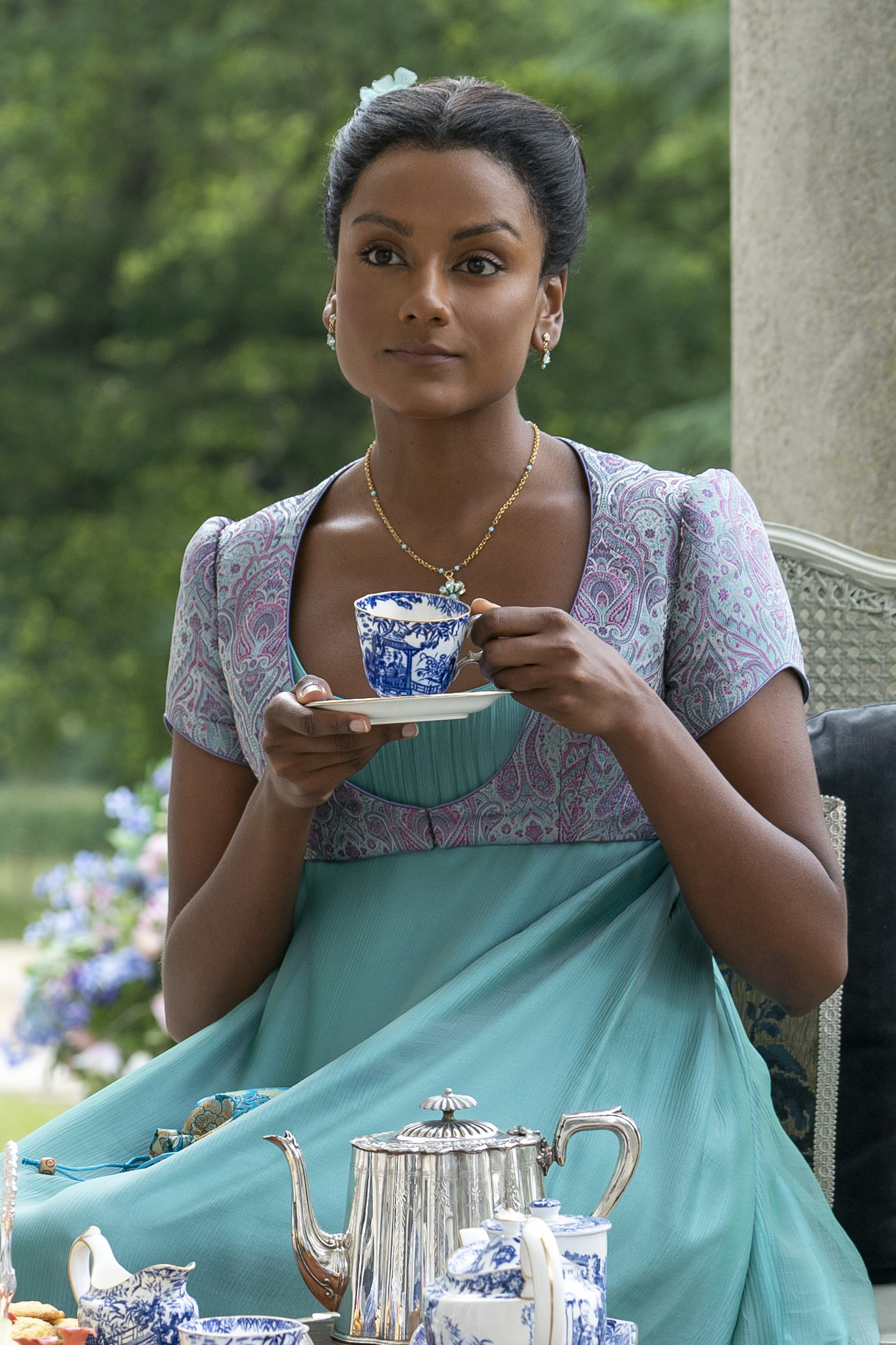Imagem mostra Simone Ashley caracterizada como Kate Sharma com vestido azul que segue a paleta de cores em tons de joias, enquanto toma chá em cena do 3º episódio da segunda temporada de Bridgerton.