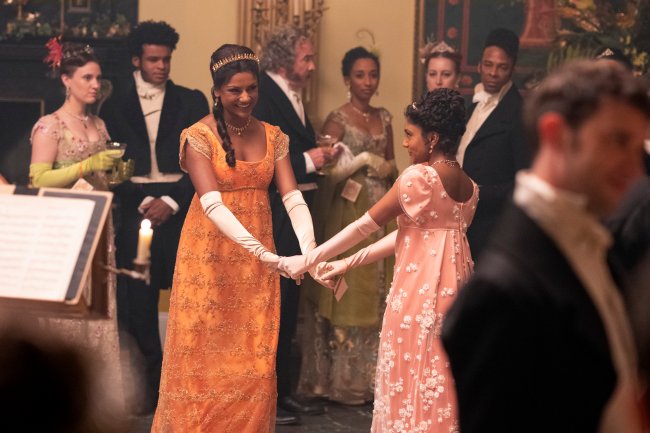 Simone Ashley como Kate Sharma e Charithra Chandran como Edwina Sharma em cena de Bridgerton; as duas estão da mãos dadas dançando em um baile no salão enquanto dão risada