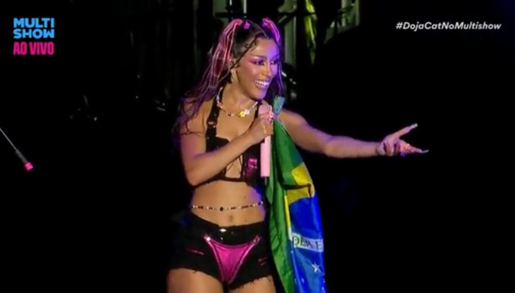 Doja Cat sorrindo enquanto segura um microfone e uma bandeira do Brasil; ela está apontando para o público que assistia seu show na sexta-feira do Lollapalooza BR 2022