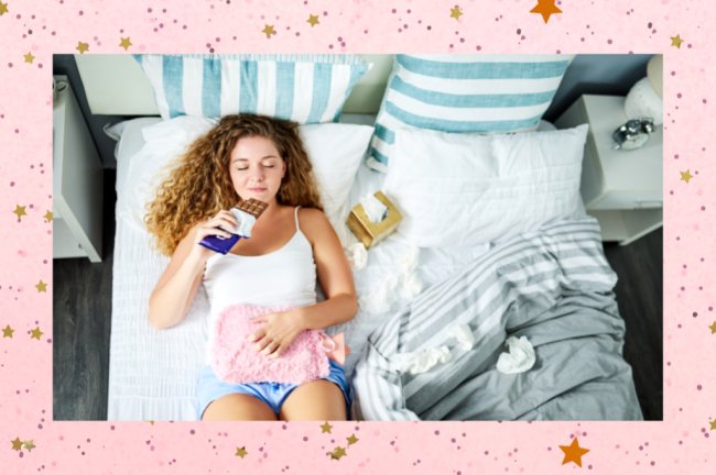 Foto de uma menina fazendo compressa com uma bolsa rosa, para cólica. Ela está deitada na cama, comendo chocolate. Ela tem cabelos claros e ondulados e pele branca.