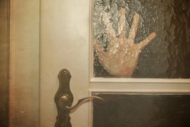 Silhueta da mão de uma mulher sobre uma porta de vidro, como se ela estivesse pedindo ajuda