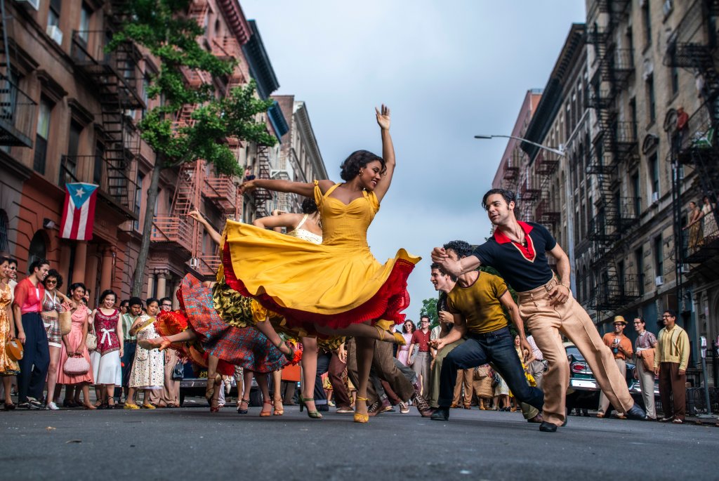Ariana DeBose como Anita e David Alvarez como Bernardo em cena de Amor, Sublime Amor; eles estão dançando na rua com outras pessoas também dançando ao fundo; ela usa um vestido amarelo com babados vermelhos na saia e ele usa uma camisa azul com detalhes em vermelho