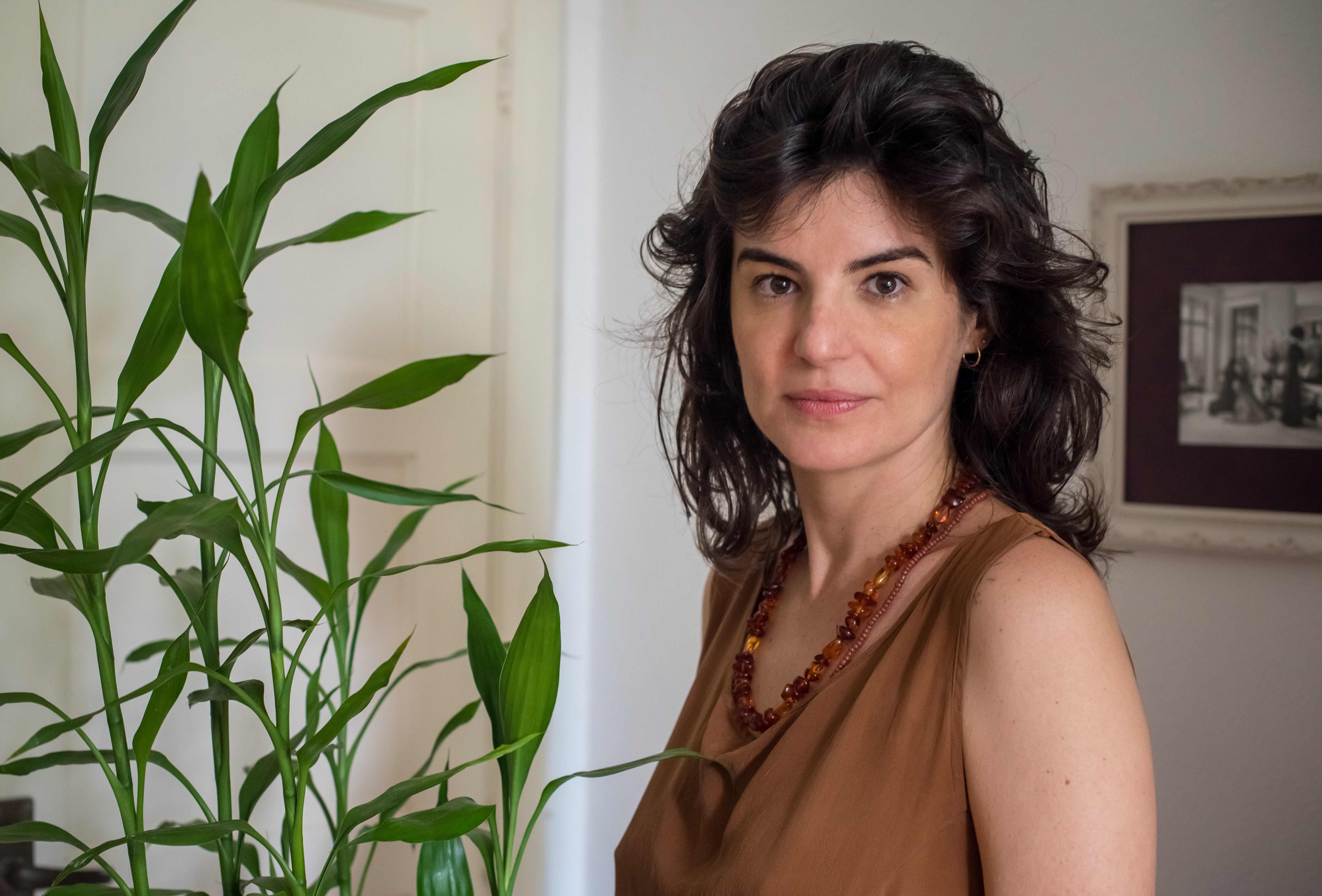 A autora Carolina Casarin olhando com expressão facial séria para a câmera, usando blusa marrom e com uma planta ao seu lado