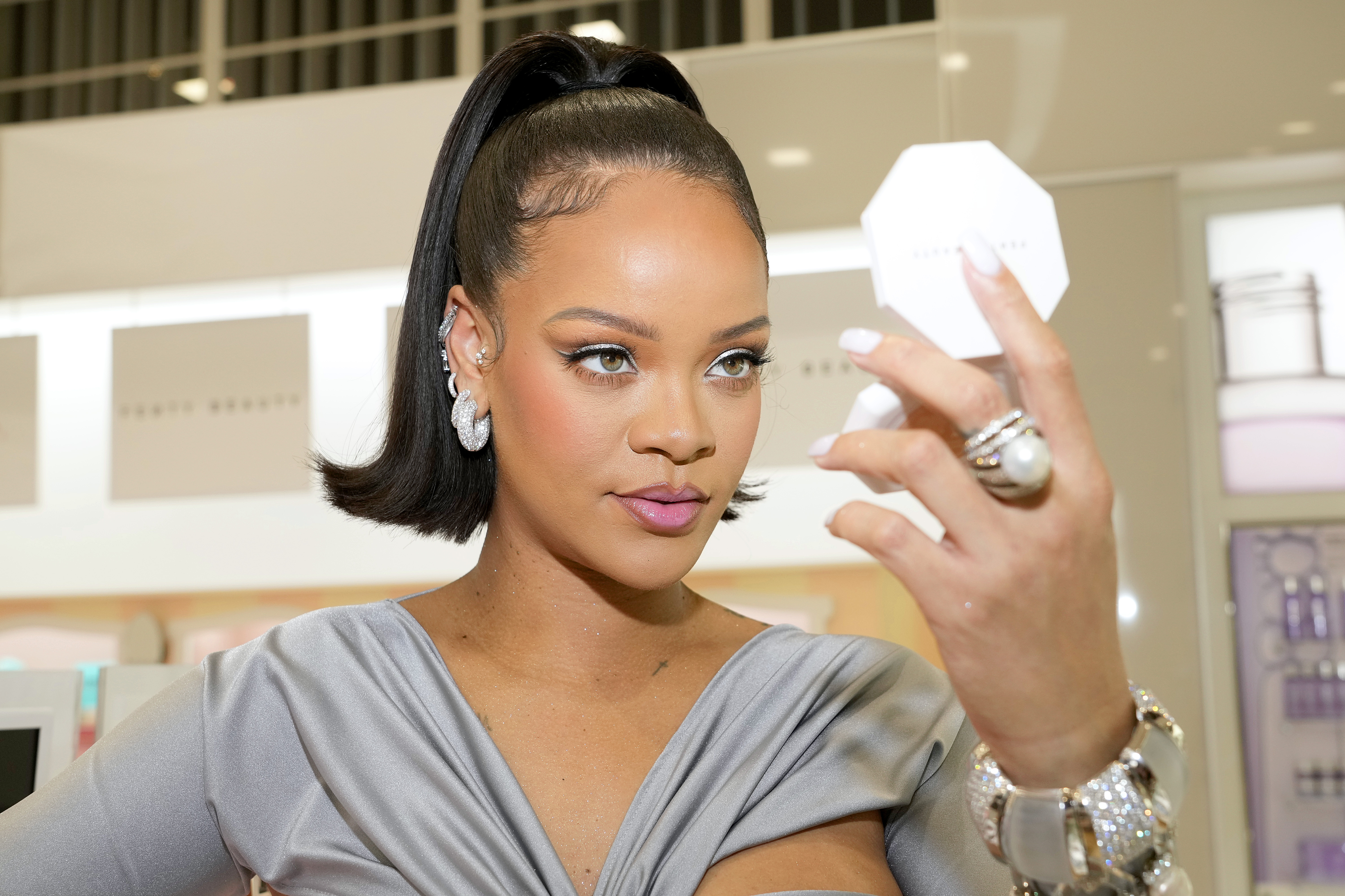 Rihanna no lançamento da Fenty Beauty na ULTA Beauty em Los Angeles, em março de 2022. Ela está com o cabelo preso em um rabo segurando um espelho de maquiagem com uma das mãos e olhando para ele