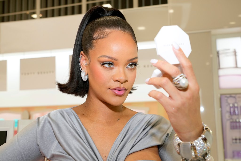 Rihanna no lançamento da Fenty Beauty na ULTA Beauty em Los Angeles, em março de 2022. Ela está com o cabelo preso em um rabo segurando um espelho de maquiagem com uma das mãos e olhando para ele