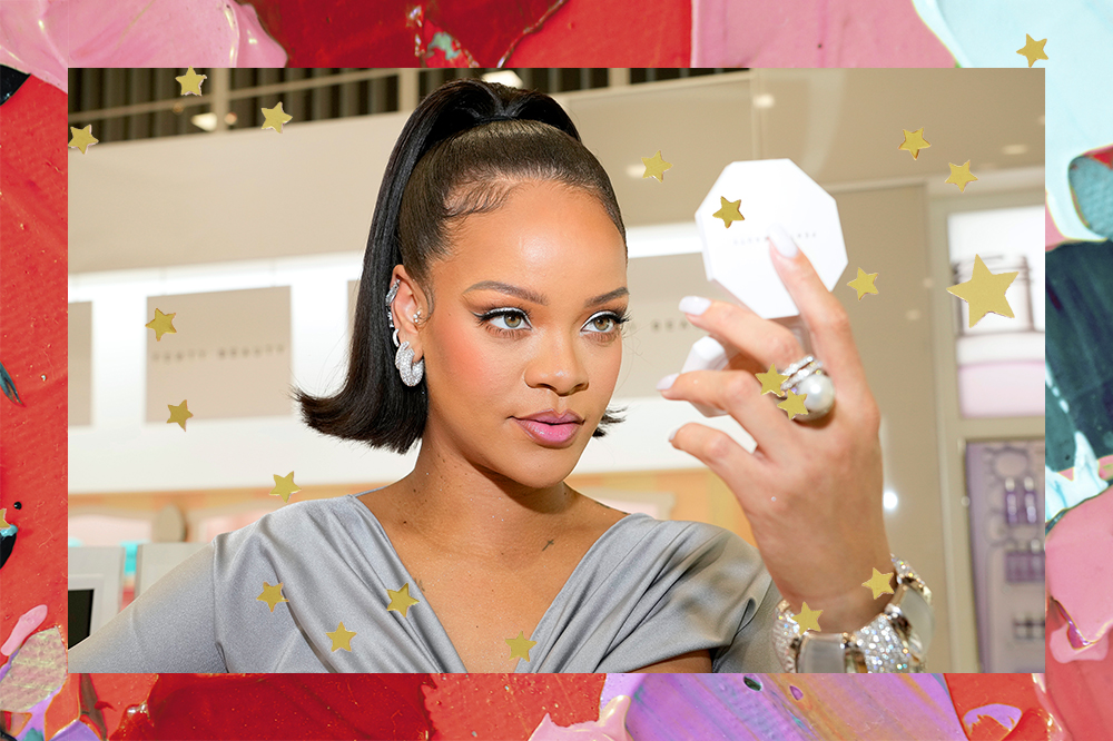 Rihanna no lançamento da Fenty Beauty na ULTA Beauty em Los Angeles, em março de 2022. Ela está com o cabelo preso em um rabo segurando um espelho de maquiagem com uma das mãos e olhando para ele. A montagem tem fundo de tintas coloridas e estrelinhas douradas