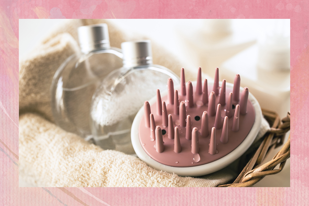 Escova massageadora de couro cabeludo rosa em uma cesta ao lado de dois frascos de vidro e montagem com fundo rosa