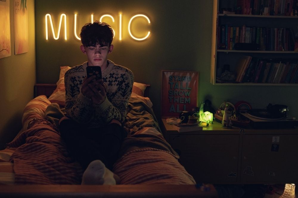 Imagem de Heartstopper, nova série da Netflix; Charlie está mexendo no celuar no sentado na cama em seu quarto com um letreiro com a palavra "MUSIC" iluminando o ambiente com uma luz amarela