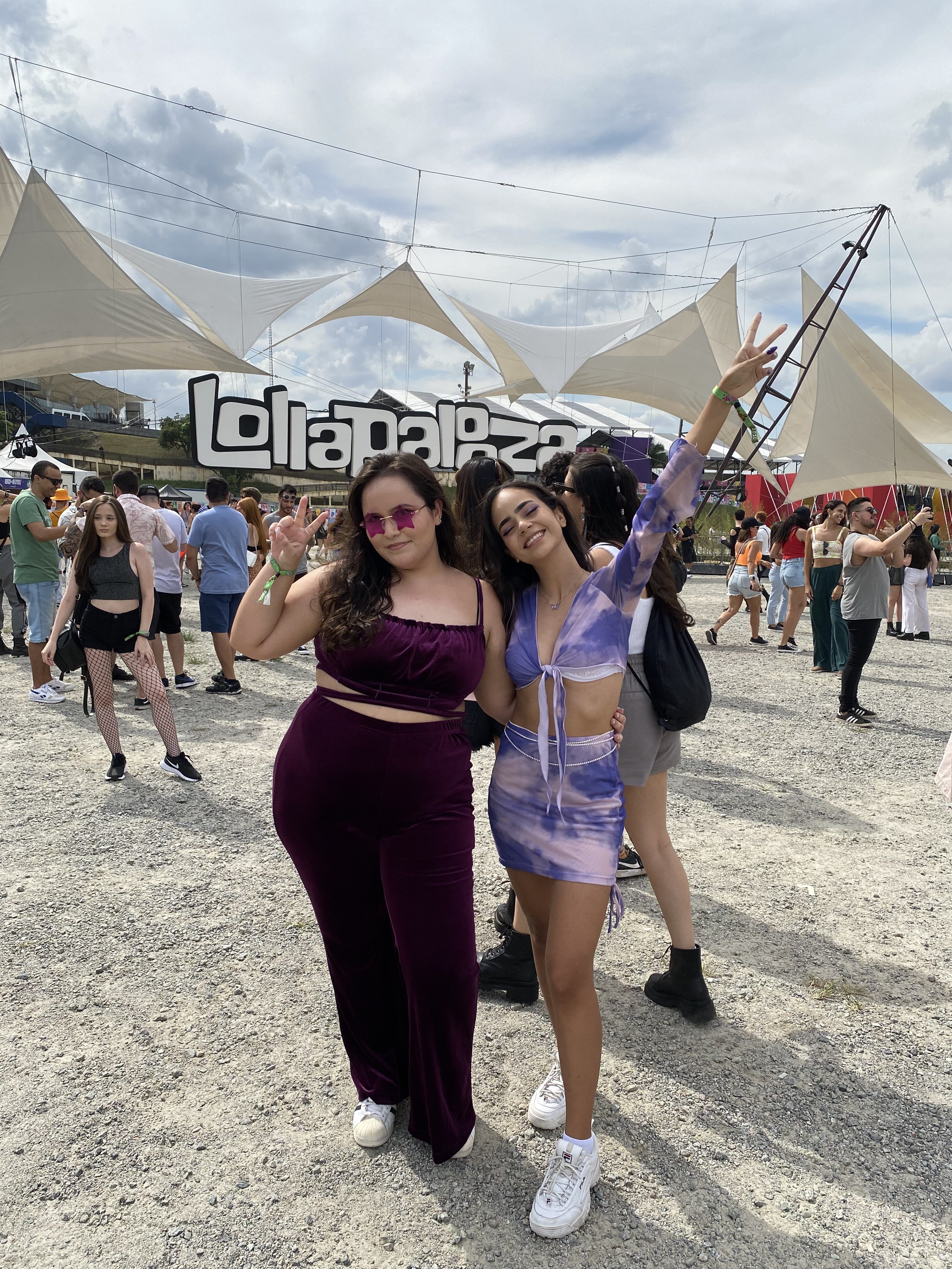 Duas garotas em frente ao letreiro do Lollapalooza usando conjuntinho. À esquerda, menina de calça e top de veludo roxo. À direita, menina de top e saia tie-dye roxo.