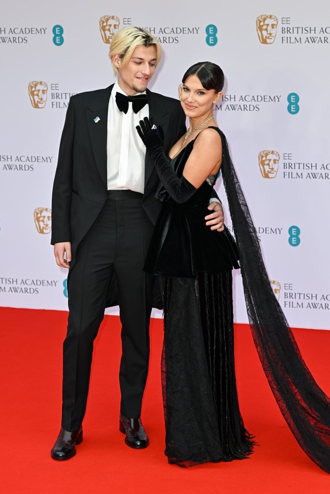 Jake Bongiovi e Millie Bobby Brown no tapete vermelho do British Academy Film Awards 2022, em Londres posando para foto e sorrindo levemente; ele está abraçando a atriz pela cintura enquanto a olha