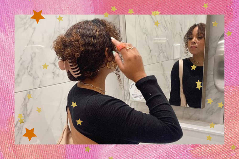 Garota de costas e de frente para espelho usando penteado com piranha de cabelo e blusa preta com uma das mãos no cabelo. O fundo da montagem é rosa com estrelinhas douradas e laranjas