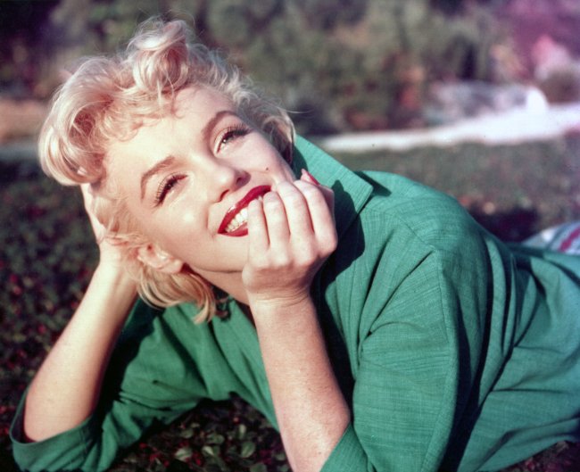 Foto da atriz Marilyn Monroe em 1954. Ela está deitada na grama da um parque, usa uma camisa verde, cabelo solto, maquiagem básica com batom vermelho. Ela olha para frente, com a mão no queixo e sorri.