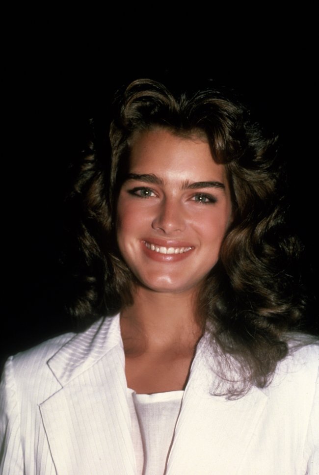 Brooke Shields nos anos 80. Ela usa uma camiseta e casaco branco, cabelo solto e maquiagem natural. Ela olha para a câmera e sorri.